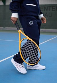 Tengerészkék tréningruha fehér részletekkel, Lacoste Teniszklub logóval. Sárga teniszütőt tart a kék pályán, fehér sportcipőt visel.