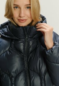 Marineblaue Steppjacke mit hohem Kragen, quergekoľteter Gestaltung und Reißverschluss, glänzender Oberfläche und gerafften Ärmeln.