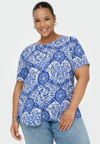 Leende kvinna med ringörhängen som bär en kortärmad topp med blå och vit blommönster och ljusblå jeans mot vit bakgrund.