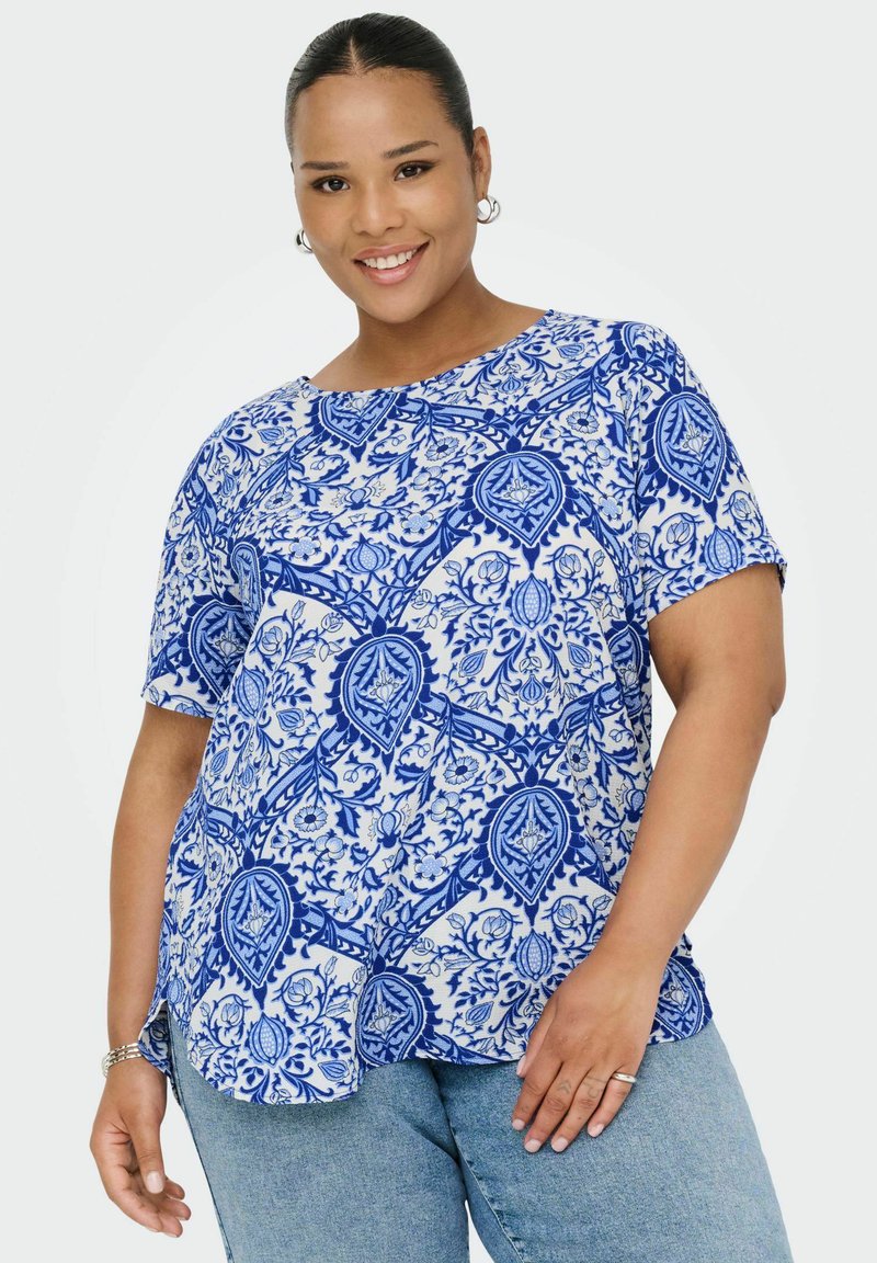 Leende kvinna med ringörhängen som bär en kortärmad topp med blå och vit blommönster och ljusblå jeans mot vit bakgrund.