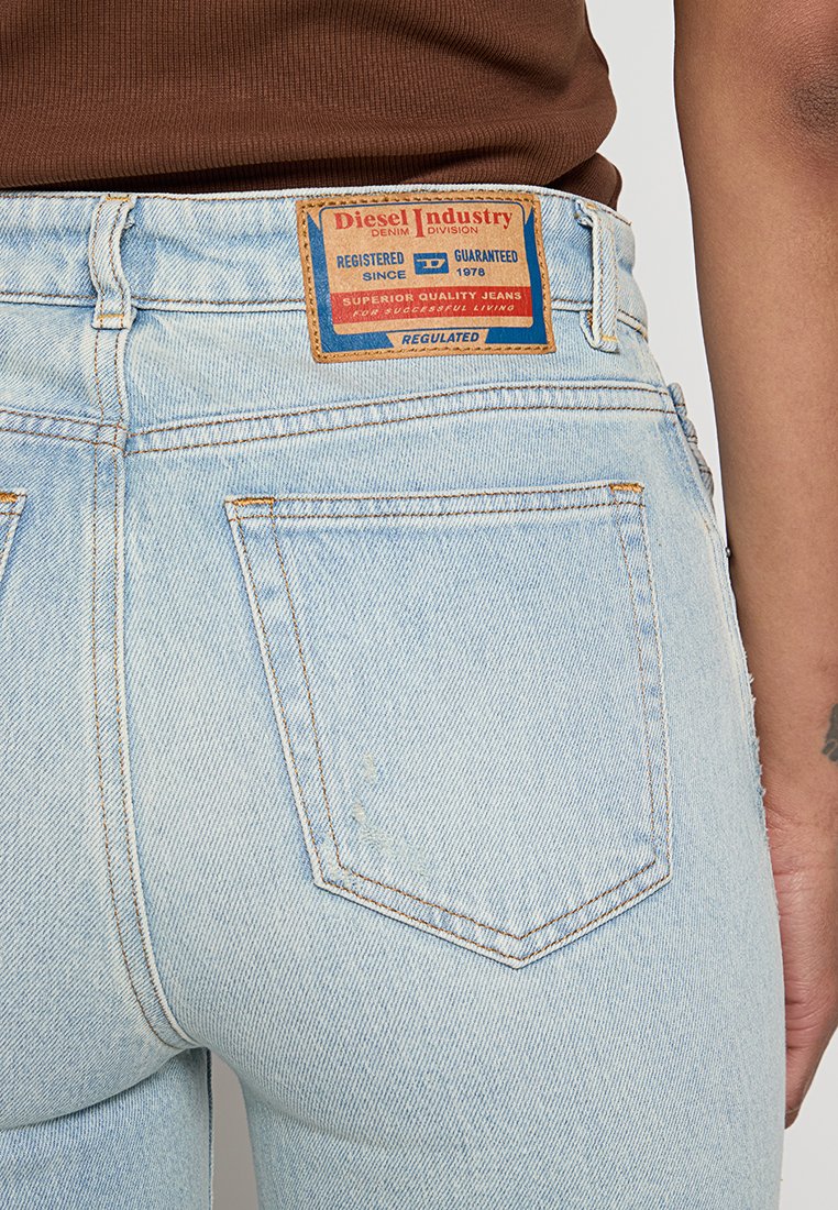 Hellblaue Jeans aus Denim mit Gesäßtasche und braunem Diesel-Industrie-Label, getragen mit einem braunen Oberteil, das einen Teil eines tätowierten Arms zeigt.