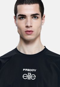 Camiseta deportiva negra con cuello redondo y mangas raglán. Tiene el texto "FREDDY elite" en blanco en el centro. Textura de tela suave.