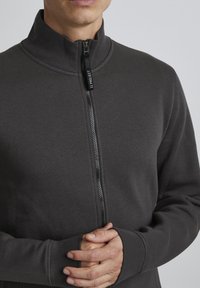 Giacca grigia con zip realizzata in tessuto morbido, caratterizzata da un collo alto e una zip con un tirante marchiato. I polsini hanno una vestibilità aderente.