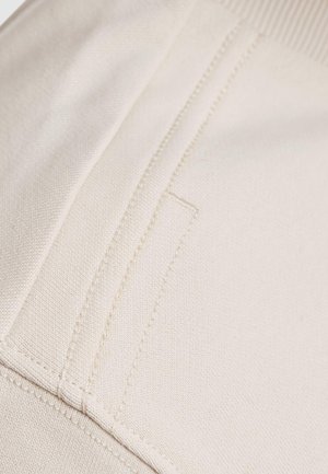 Tessuto di cotone beige con cuciture visibili e una superficie piatta e liscia. Presenta un dettagliato tasca discreta lungo la cucitura.