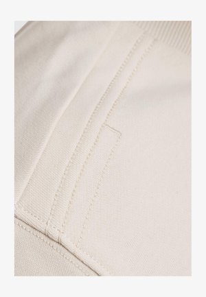 Tessuto di cotone beige con cuciture visibili e una superficie piatta e liscia. Presenta un dettagliato tasca discreta lungo la cucitura.