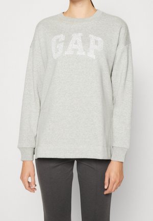 Henkilö pukeutunut vaaleanharmaaseen pitkähihaiseen collegepaitaan, jossa on "GAP"-logo rinnalla, ja tummanharmaisiin housuihin, seisoo valkoista taustaa vasten.