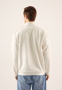 Balts merserizētas vilnas džemperis ar augstu apkakli, izceļot smalkas šūšanas detaļas un ribotās aproces, valkāts ar gaiši zilām džinsa biksēm.