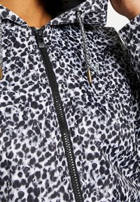 Veste à imprimé léopard noir et blanc avec fermeture éclair noire, capuche, et cordons noirs et blancs à embouts dorés.