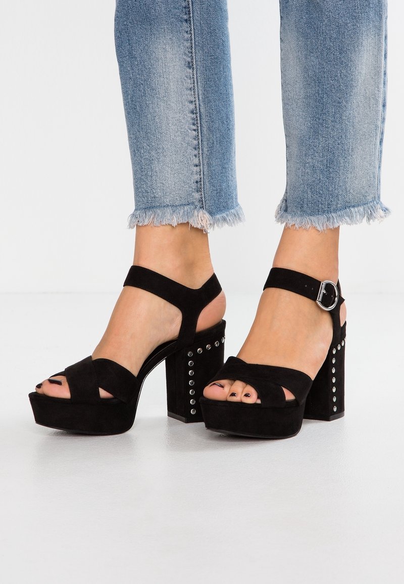 Pieds portant des sandales compensées en daim noir avec des brides à la cheville et des clous argentés sur des talons épais, associées à un jean bleu à ourlet effiloché.