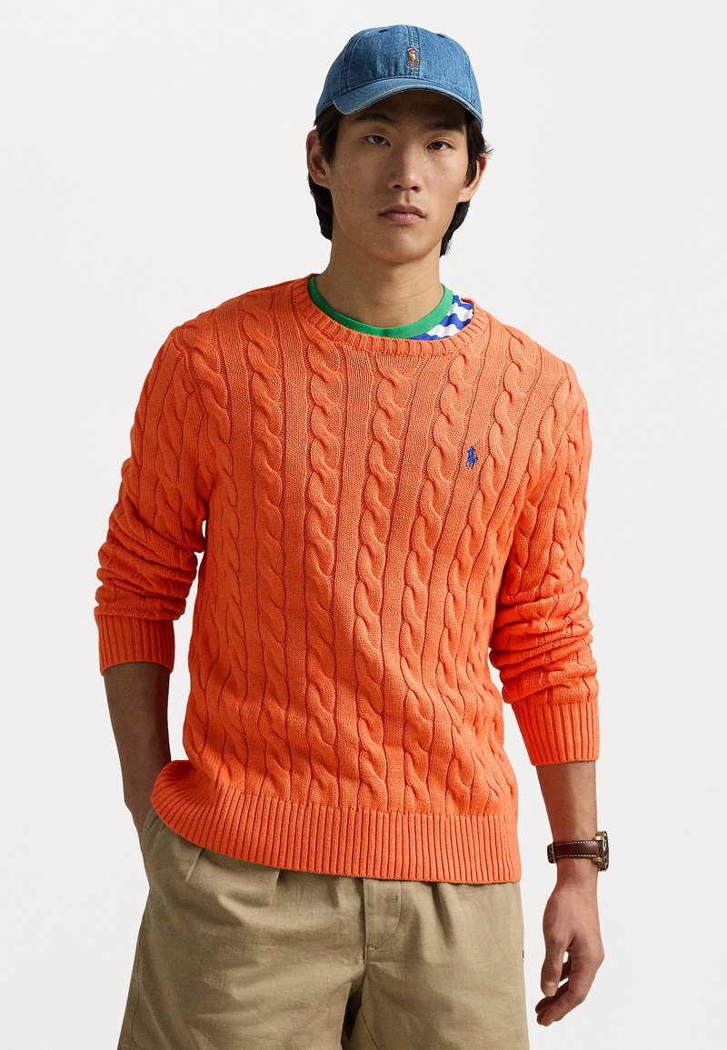Orange cable-knit sweater with ribbed cuffs and hem. Worn over a green shirt featuring a striped pattern. Blue denim cap with logo.