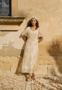 Femme en robe longue légère à fleurs et chaussures tissées, debout contre un mur en pierre beige texturé sous un rebord de fenêtre dans une lumière douce.