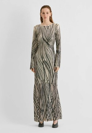 Un mannequin se tient devant un fond gris uni, vêtu d’une longue robe crème à larges rayures géométriques noires et de chaussures noires.
