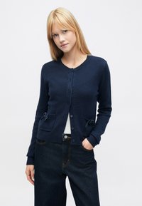 Sötétkék kardigán texturált kötött anyagból, kerek nyakkivágással, gombos lezárással, két elülső zsebbel masnival és bordázott mandzsettákkal.