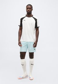 Άνδρας αθλητής που φοράει λευκό και μαύρο αθλητικό μπλουζάκι Puma, ανοιχτό μπλε σορτς, λευκές κάλτσες Puma μέχρι το γόνατο και λευκά παπούτσια με πολύχρωμες σόλες, όρθιος.