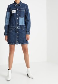 Denim shirt dress featuring a mix of light and dark blue patches, button front, point collar, chest pocket, and side pockets. White ankle boots.