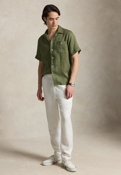 Jeune homme portant une chemise à manches courtes boutonnée couleur vert olive, un pantalon blanc, des baskets blanches et une montre-bracelet marron, debout sur un fond neutre.