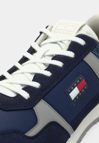 Marineblauwe sneakers met suede en stoffen panelen, witte veters en een grijze accent; heeft een logopatch in rood, wit en blauw.