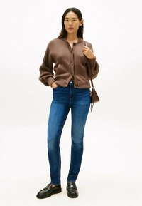 Vrouw staat in een bruine knopen cardigan, blauwe skinny jeans, zwarte instappers en een bruine schoudertas tegen een egale achtergrond.