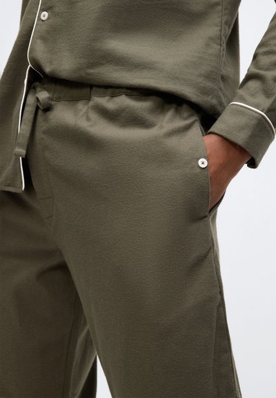 Close-up van een persoon die olijfgroene broek en bijpassend geknoopte shirt met witte bies draagt, hand in de pocket.