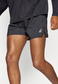 Shorts de sport noirs avec un motif texturé, présentant un ourlet fendu sur le côté, des détails de logo argenté et des accents rouges sur la ceinture.