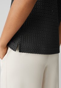 Top nero con texture e piccole aperture, con orlo dritto e spacchetti laterali, abbinato a pantaloni color crema.