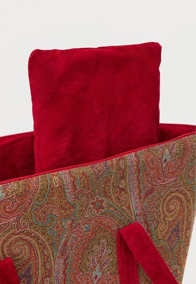 Large red suede pillow partially inside a paisley-patterned fabric bag with red suede handles against a plain background.