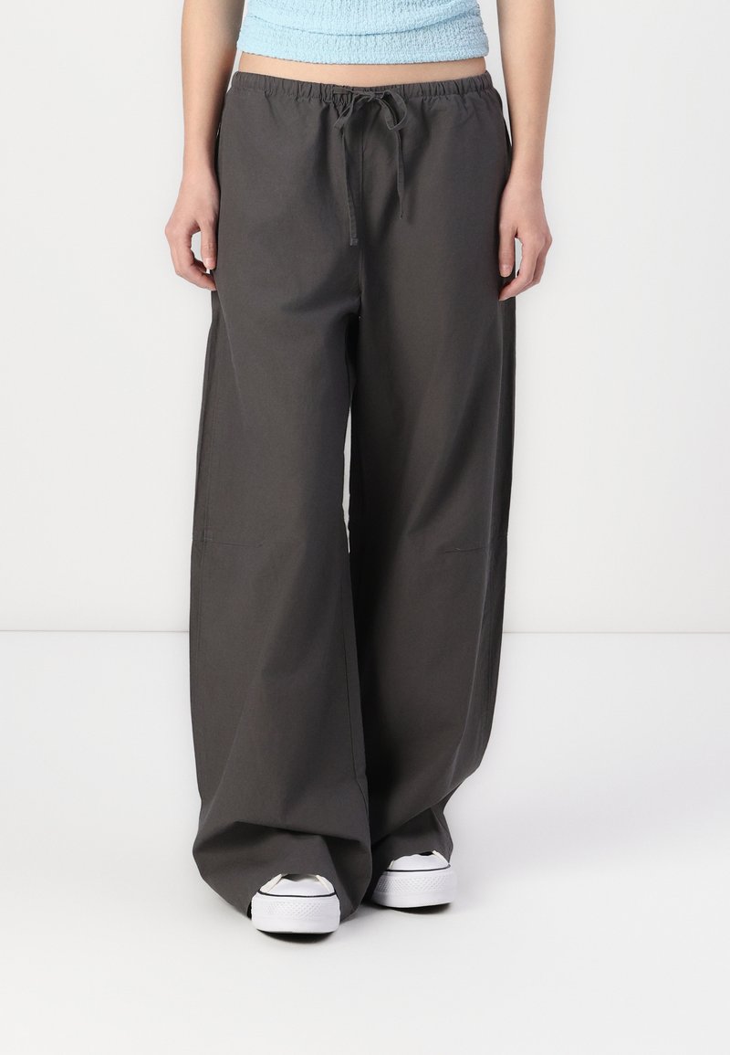 Pantaloni larghi di colore grigio scuro con vita regolabile, realizzati in tessuto morbido. Presentano due tasche laterali e una vestibilità comoda. Abbinati a sneakers bianche.