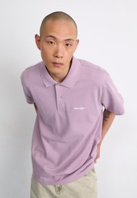Polo lilas avec un col et trois boutons, arborant un logo blanc. Le tissu semble lisse, avec une coupe décontractée et des manches courtes.