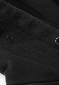 Svart skjorte med knapper laget av mykt, teksturert stoff, med en subtil brodert logo og dekorativ detalj i sirkulær metallknapp.