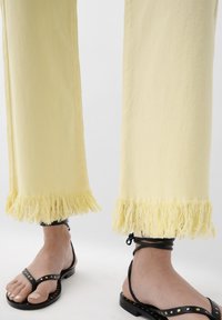 Pantaloni din denim galbeni cu margini franjurate, asortați cu sandale plate negre cu știfturi aurii. Prim-plan al picioarelor și încălțămintei.