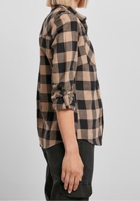 Checkered flannel shirt in beige and black, with a buttoned roll-tab sleeve, front pocket, and a straight hem. Material appears soft and textured.