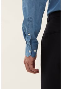 Chemise en denim bleu clair à manches longues avec des poignets boutonnés et une texture lisse, portée avec un pantalon sombre. Les poignets présentent des boutons blancs.