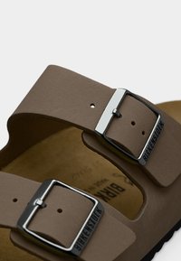 Κοντινό πλάνο από καφέ δερμάτινα λουριά σανδαλιών με ασημένιες μεταλλικές πόρπες που φέρουν ανάγλυφο το κείμενο "BIRKENSTOCK".