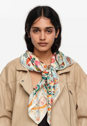 Mujer de cabello oscuro que lleva una chaqueta beige y una bufanda de seda colorida con un estampado atada alrededor de su cuello, con un fondo liso.