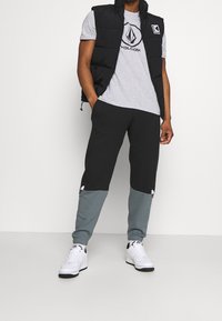 Svart vadderad väst, grå T-shirt med logotyp, svarta byxor med teal-färgade muddar och vita sneakers. Funktioner inkluderar en väst med dragkedja och logotypdetaljer.