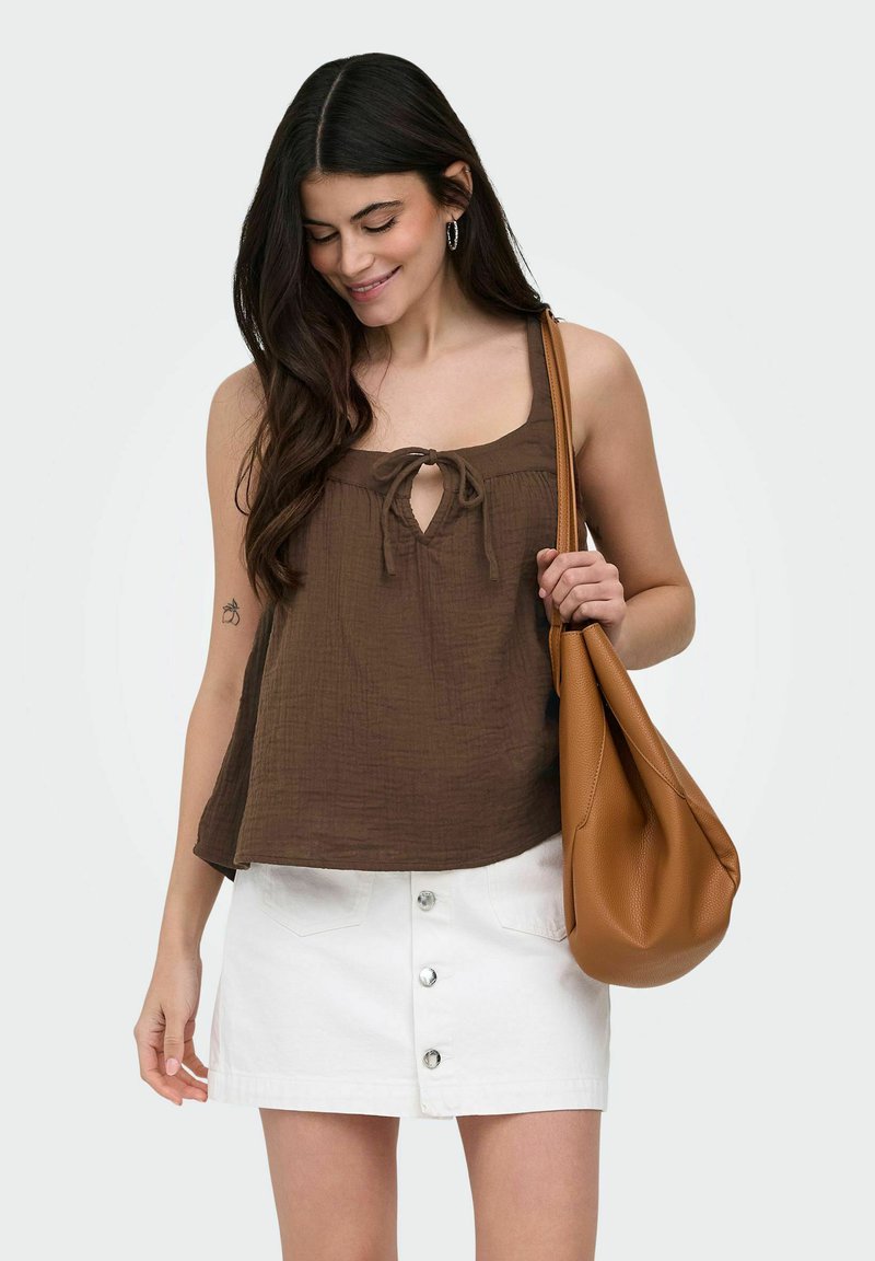 Jeune femme portant un haut marron sans manches et une jupe blanche boutonnée devant, tenant un grand sac à bandoulière en cuir marron.