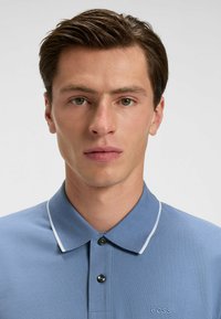 Ljusblå polotröja i texturerad tyg, med klassisk krage, vit kantning och en broderad logotyp på bröstet.