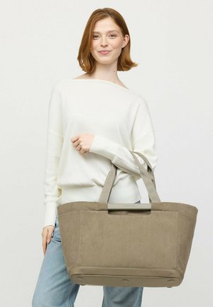 Jonge vrouw in witte trui en spijkerbroek die een grote beige tas met dubbele hengsels vasthoudt tegen een effen achtergrond.