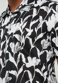 Svart och vit abstrakt skjorta med bladmönster och korta ärmar på en person med medelhudton.
