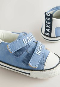 Zilā suede kedas ar baltajiem akcentiem, Velcro jostām un teksturētiem zoliņām. Iespējas ir zīmola burti uz jostām un kontrastējoša papēža cilpa.
