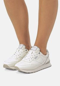 Weiße Sneakers mit einem glatten Lederobermaterial, strukturierten Akzenten und einer gepolsterten Sohle. Merkmale sind silberne Details und ein perforiertes Zehen-Design.