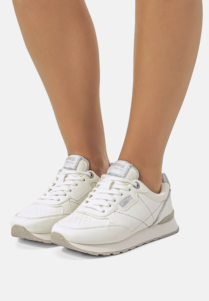 Weiße Sneakers mit einem glatten Lederobermaterial, strukturierten Akzenten und einer gepolsterten Sohle. Merkmale sind silberne Details und ein perforiertes Zehen-Design.