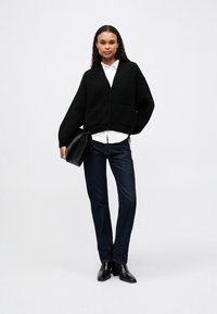 Svart strikket cardigan med V-hals og to lommer, brukt over en hvit skjorte, kombinert med mørke denimjeans og svarte ankelstøvler.