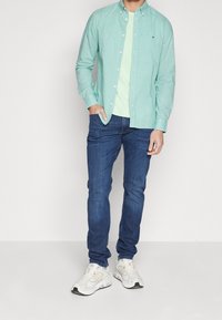 Ljusgrön rutad skjorta över en myntgrön T-shirt, tillsammans med blå jeans och vita sneakers, på en neutral bakgrund.