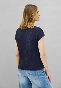 Top blu navy a maniche corte realizzato in un tessuto testurizzato con un motivo ondulato, abbinato a jeans larghi di colore blu chiaro.