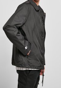 Personne portant une veste noire boutonnée avec des poches latérales et un logo étoile blanc sur la manche gauche, associée à un pantalon foncé et une chemise rayée en dessous.