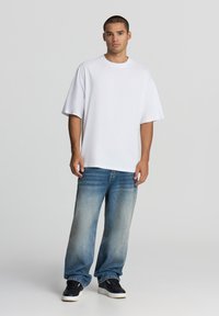 Biała, oversize bawełniana koszulka z krótkim rękawem, w połączeniu z wyblakłymi niebieskimi dżinsami i czarnymi sneakersami, stojąca na neutralnym tle.