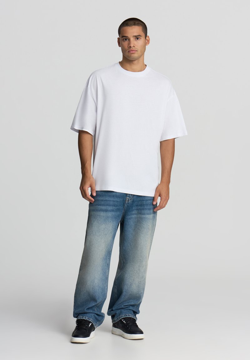 Biała, oversize bawełniana koszulka z krótkim rękawem, w połączeniu z wyblakłymi niebieskimi dżinsami i czarnymi sneakersami, stojąca na neutralnym tle.