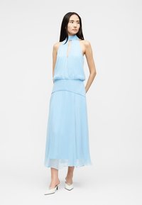 Γυναίκα με αμάνικο ανοιχτό γαλάζιο φόρεμα halter με ντεκολτέ κλειδιού και ελαφρώς ροϊκή μίντι φούστα, φοράει λευκά παπούτσια με τακούνι, στέκεται μπροστά σε μονόχρωμο φόντο.