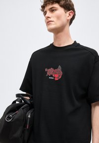 Νέος άνδρας που φοράει μαύρο μπλουζάκι με κόκκινο γραφικό τριαντάφυλλο και το κείμενο "HUGO", κρατώντας μια μαύρη τσάντα duffel σε ανοιχτό φόντο.